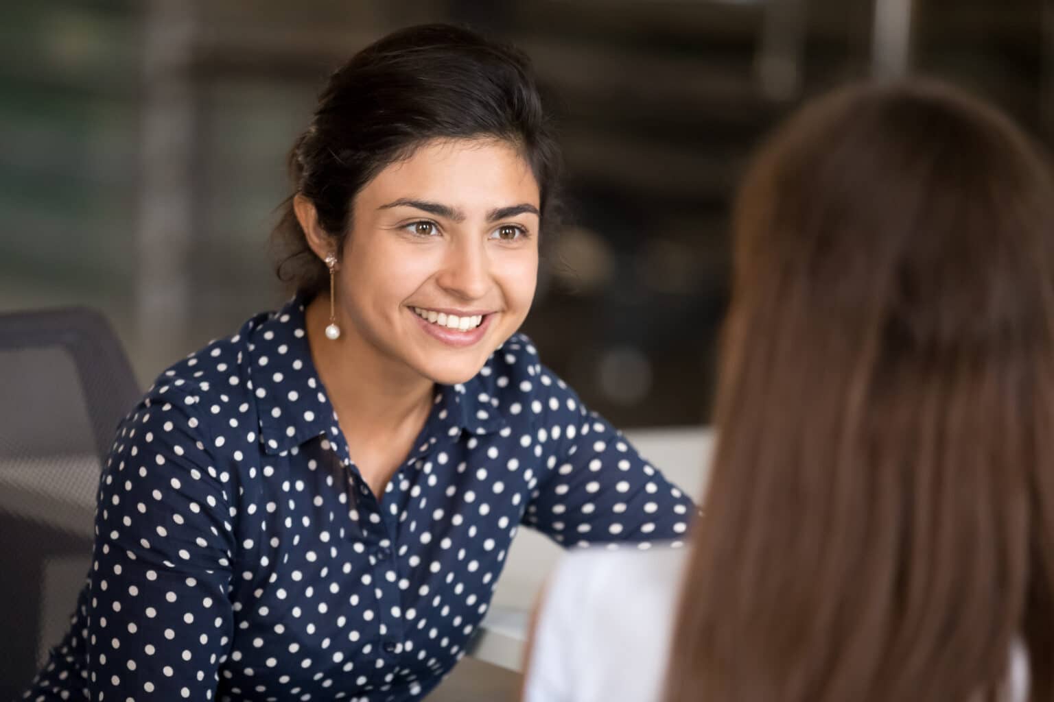 Indian attractive positive woman talking with colleague sitting at desk in office room, focus on hindu female. Diverse millennial smiling employees have break communicating together at coworking area